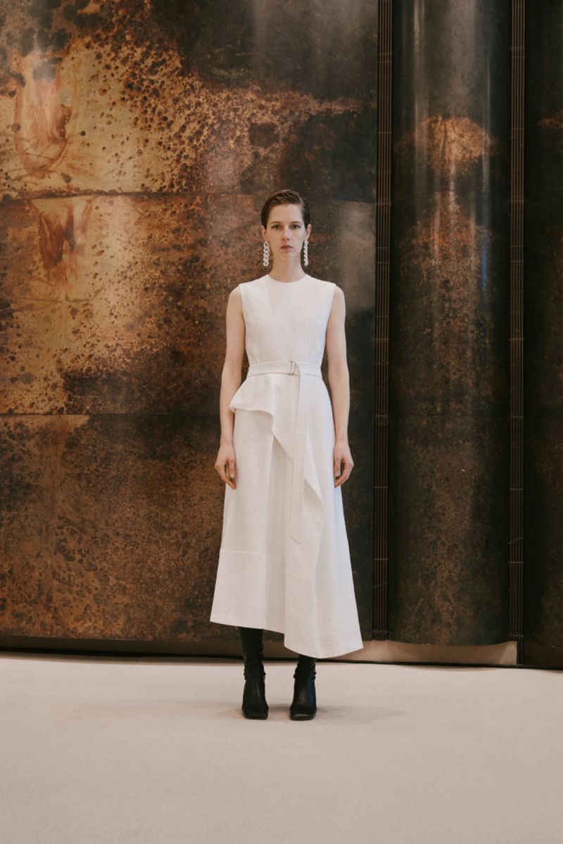 Model standing in a sleeveless white dress with an asymmetric hem against a dark textured metallic wall.