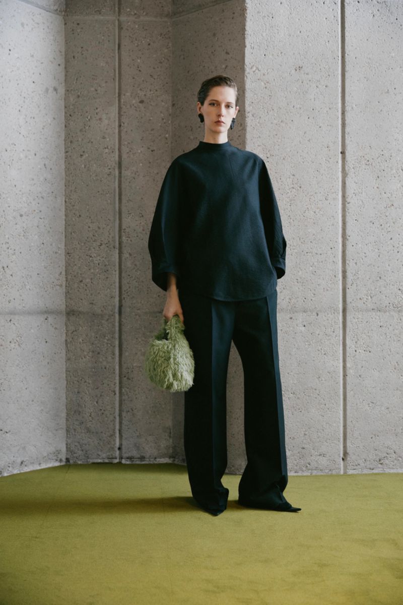 Studio shot of a model in a dark teal outfit and green handbag. The model is standing in front of a grey wall with green carpet.