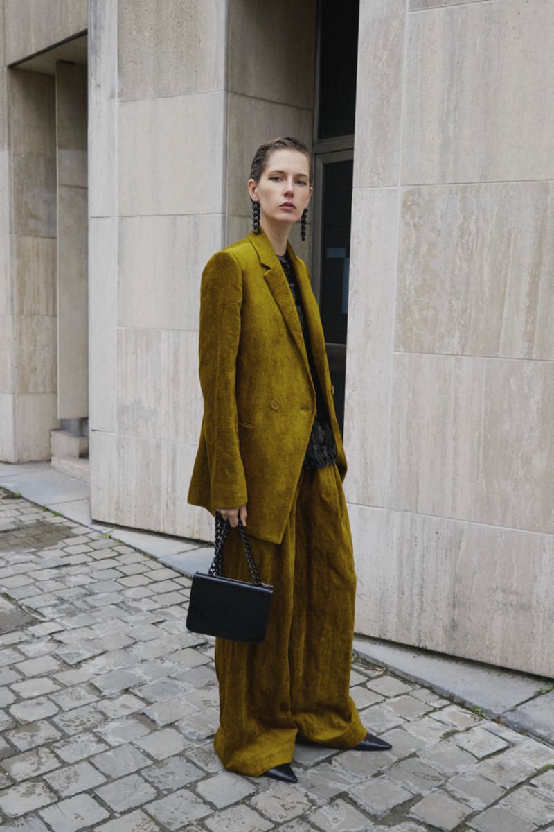 Stylish adult woman in an olive green suit stands on a cobblestone street with a black purse in an outdoor urban setting.