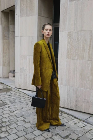 Stylish adult woman in an olive green suit stands on a cobblestone street with a black purse in an outdoor urban setting.