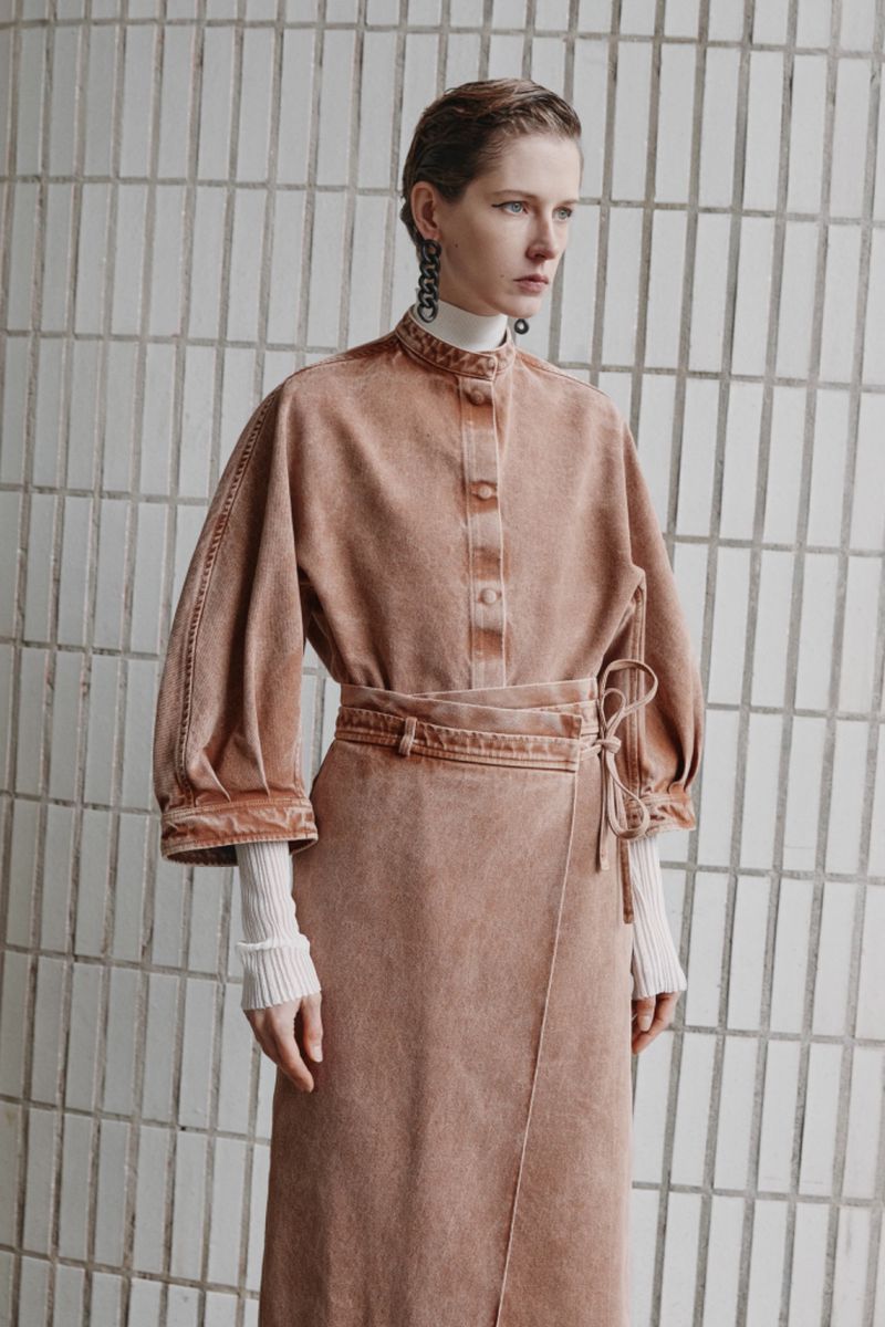 A woman in an old-rose, long-sleeved, midi-length dress with matching belt stands against a white-tiled wall in a medium indoor shot.