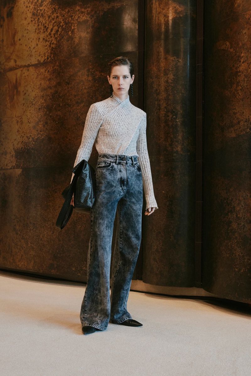 A slender woman poses indoors wearing a ribbed sweater, acid-wash jeans, and heels while carrying a black purse over her shoulder.