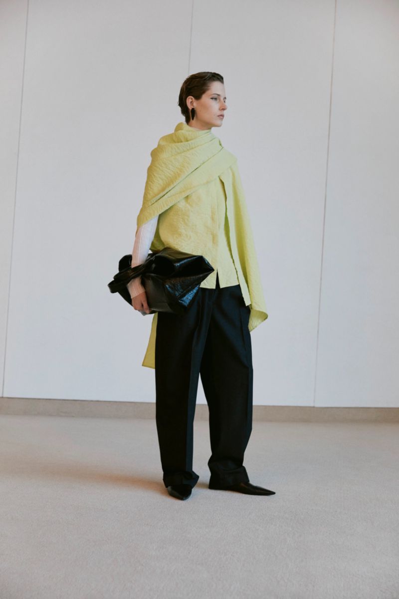 Fashion shot of an adult woman wearing a pale green top, draped shawl, black pants and holding a black bag inside a studio setting.