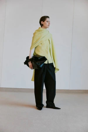 Fashion shot of an adult woman wearing a pale green top, draped shawl, black pants and holding a black bag inside a studio setting.