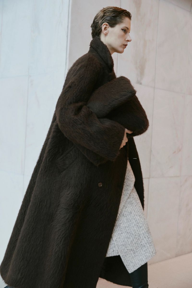 A woman in a brown coat walks in a minimalistic indoor setting, carrying a soft clutch, with a clean and modern aesthetic.