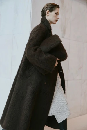 A woman in a brown coat walks in a minimalistic indoor setting, carrying a soft clutch, with a clean and modern aesthetic.