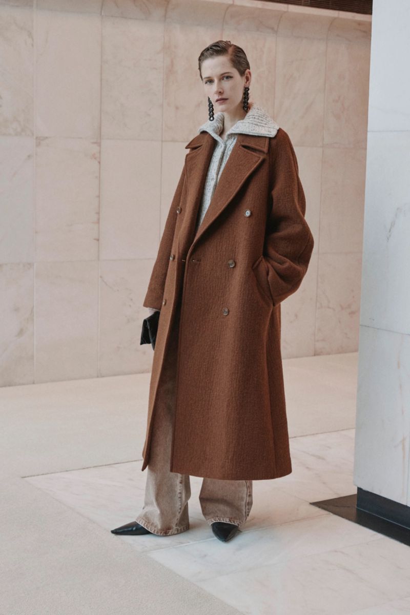 Model wearing a long brown double-breasted coat and patterned knit collar against a polished stone wall interior.