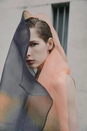 Close-up portrait of a fair-skinned adult woman wearing a sheer, peach-colored garment that is partially obscured by a sheer, blue fabric.