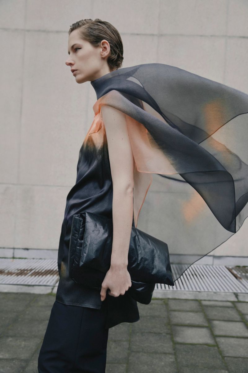 Fashion shoot featuring a female model with short hair wearing a tunic and flowing scarf, holding a black clutch, posing outdoors.