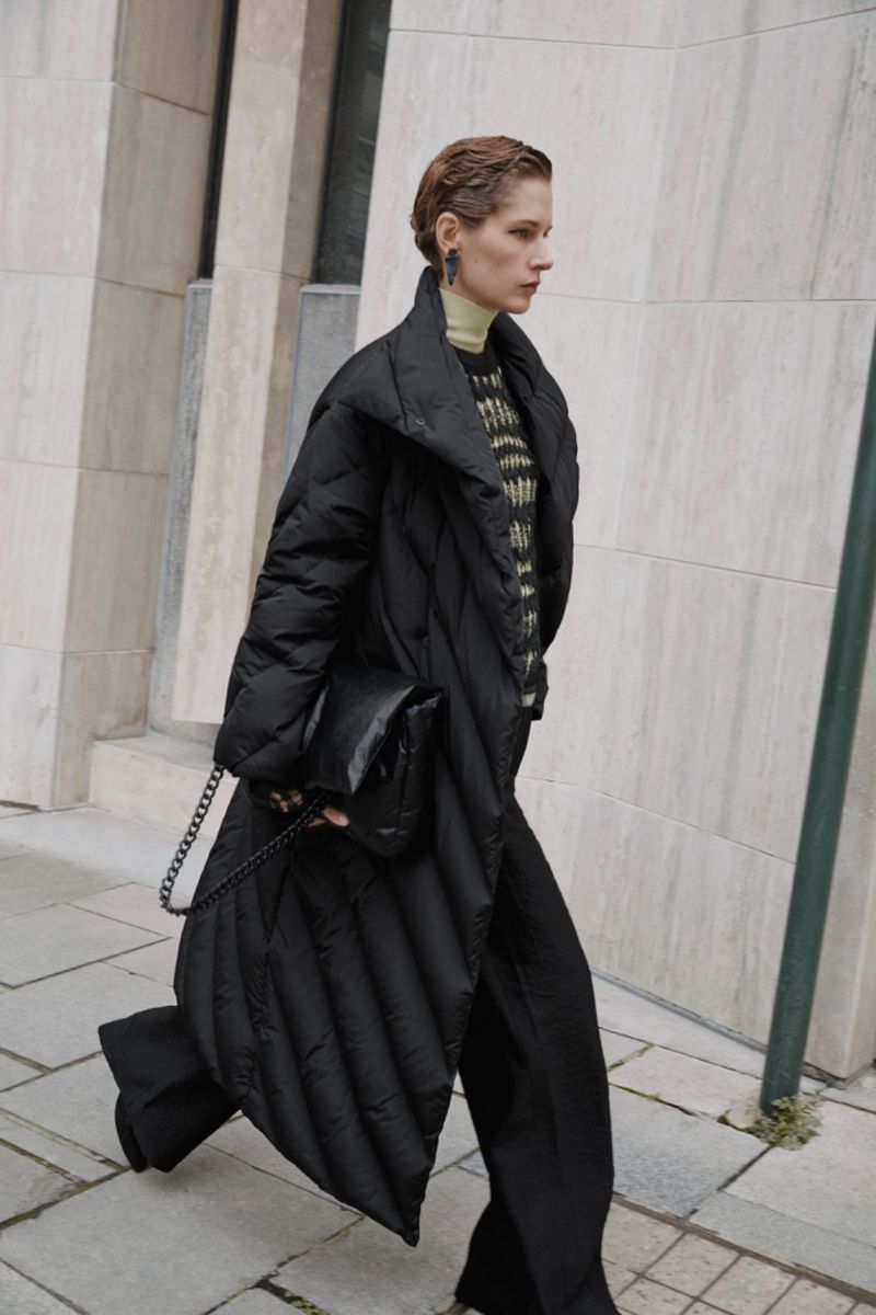 Fashion shot of a woman in a black puffer coat and black trousers walking outdoors with a black purse on a city sidewalk.