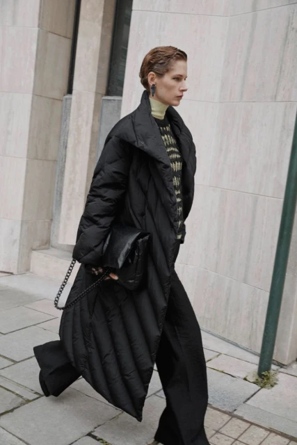 Fashion shot of a woman in a black puffer coat and black trousers walking outdoors with a black purse on a city sidewalk.