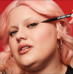 Close-up beauty portrait of a model with pink hair applying black liquid eyeliner against a red background.