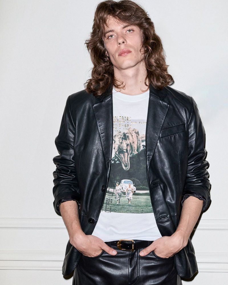 Adult male with long hair wearing a graphic T-shirt and leather blazer and pants, posing against a plain white background indoors.