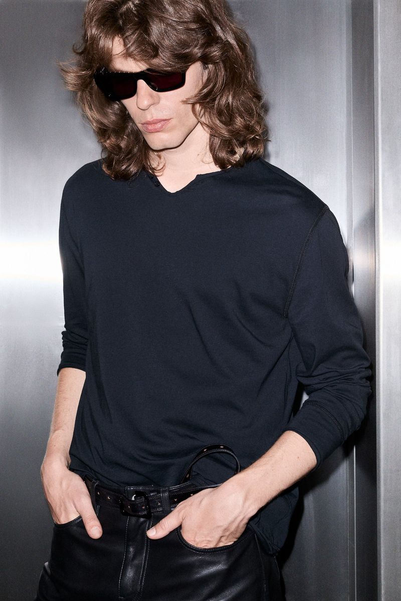 A brunette male model with sunglasses poses in a long-sleeved shirt and leather pants against a textured, silver background.