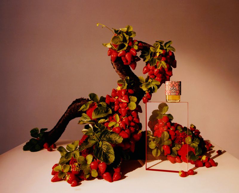 Perfume bottle with floral cap atop a clear platform, next to a sculpture of strawberries and leafy green vines on a white surface.