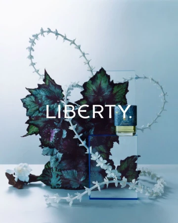 Studio shot of a Liberty perfume bottle displayed on a clear stand, surrounded by dark green leaves and small white flower decorations.