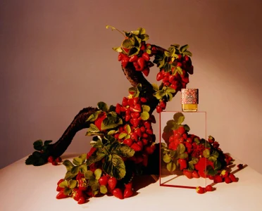 Perfume bottle resting on a glass stand next to an ornate twisting branch laden with strawberries and green leaves.
