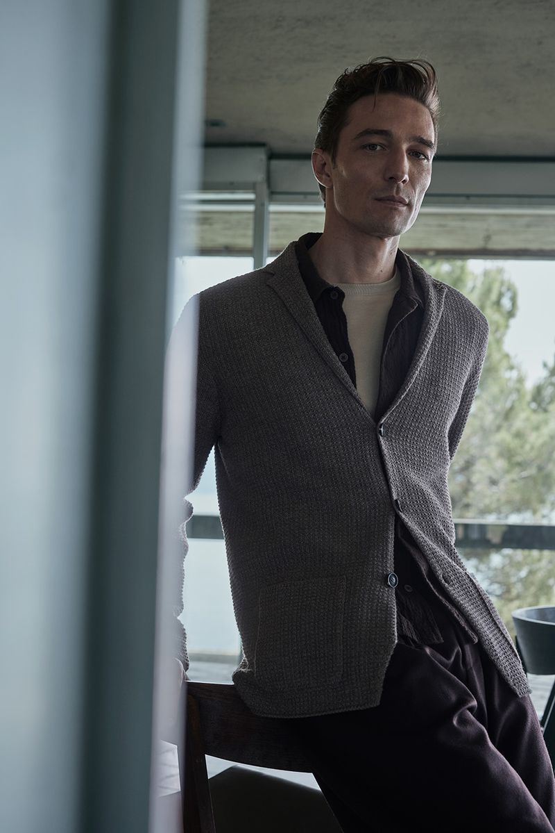 Adult man wearing brown cardigan and dress pants is posing indoors with natural light for fashion editorial photography.