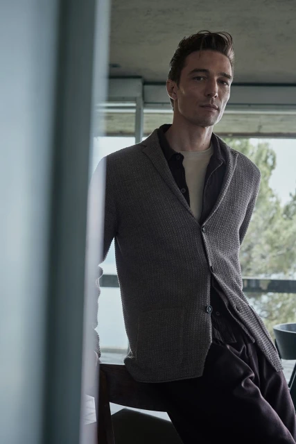 Adult man wearing brown cardigan and dress pants is posing indoors with natural light for fashion editorial photography.