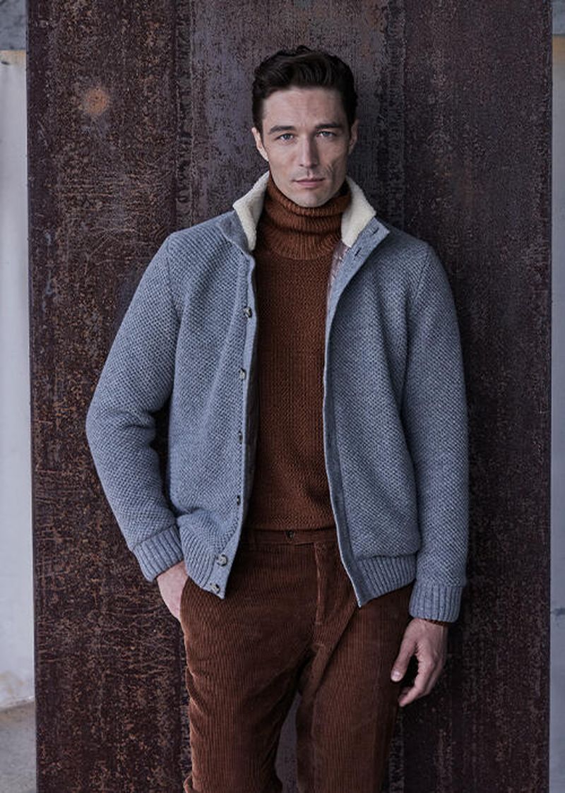 Adult male in gray wool cardigan with tan turtleneck and corduroy pants against a textured wall in a medium studio shot.
