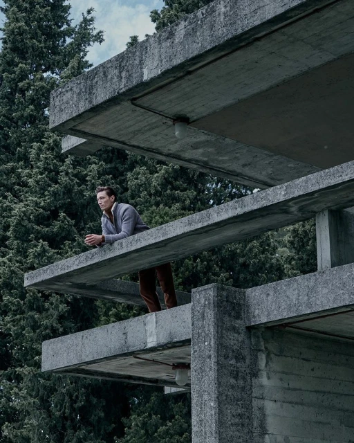 Adult male poses on a modern concrete architectural structure, set against a backdrop of lush green trees and cloudy skies.