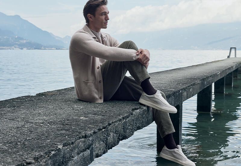 Man seated on a concrete pier overlooking a lake with mountains in the background, wearing a knit cardigan and trousers.