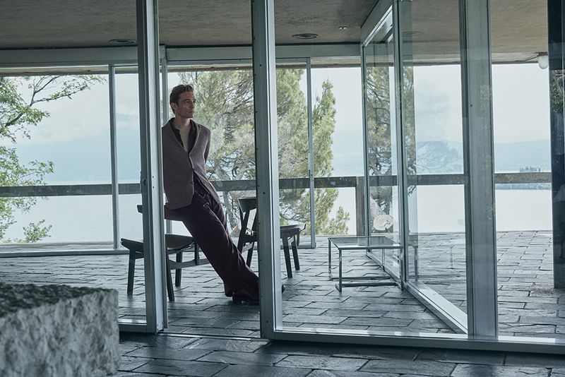 Adult male wearing a vest and pants leans against the aluminum frame of a glass wall in a modern building, overlooking a lake.