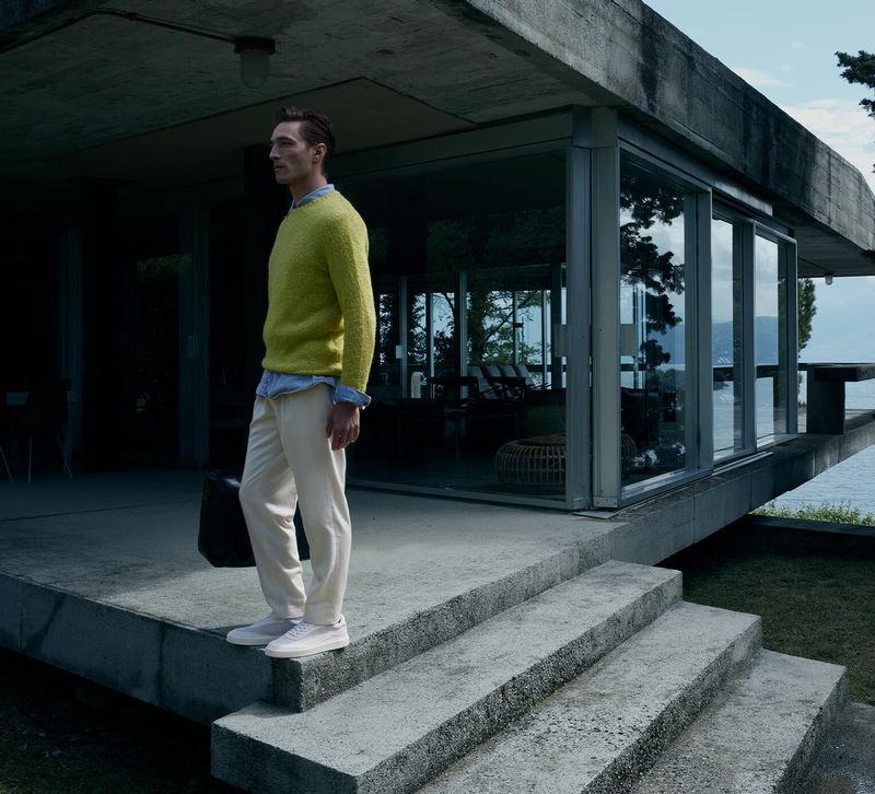 Fashionable man in a yellow sweater stands on concrete steps outside a modern house with views of a lake on a cloudy day.