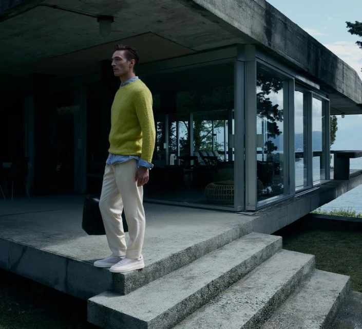 Fashionable man in a yellow sweater stands on concrete steps outside a modern house with views of a lake on a cloudy day.