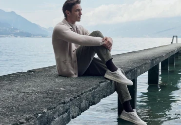 Man seated on a concrete pier overlooking a lake with mountains in the background, wearing a knit cardigan and trousers.