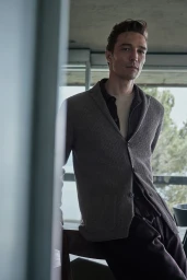 Portrait of a male model wearing a grey knitted cardigan and dark trousers indoors against a window background.