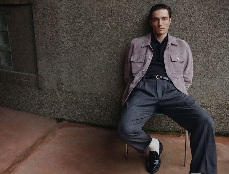 Adult male seated wearing a grey suit with a lavender jacket, shiny black loafers, and white socks, posing against a textured wall.