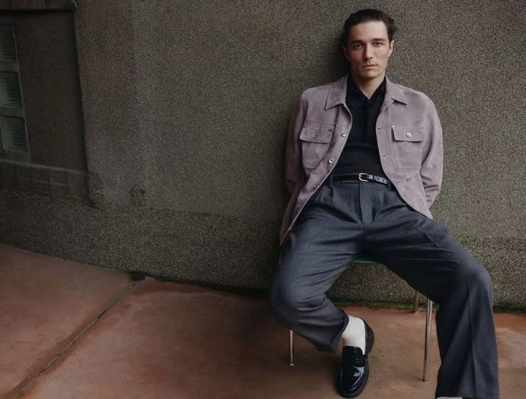 Adult male seated wearing a grey suit with a lavender jacket, shiny black loafers, and white socks, posing against a textured wall.