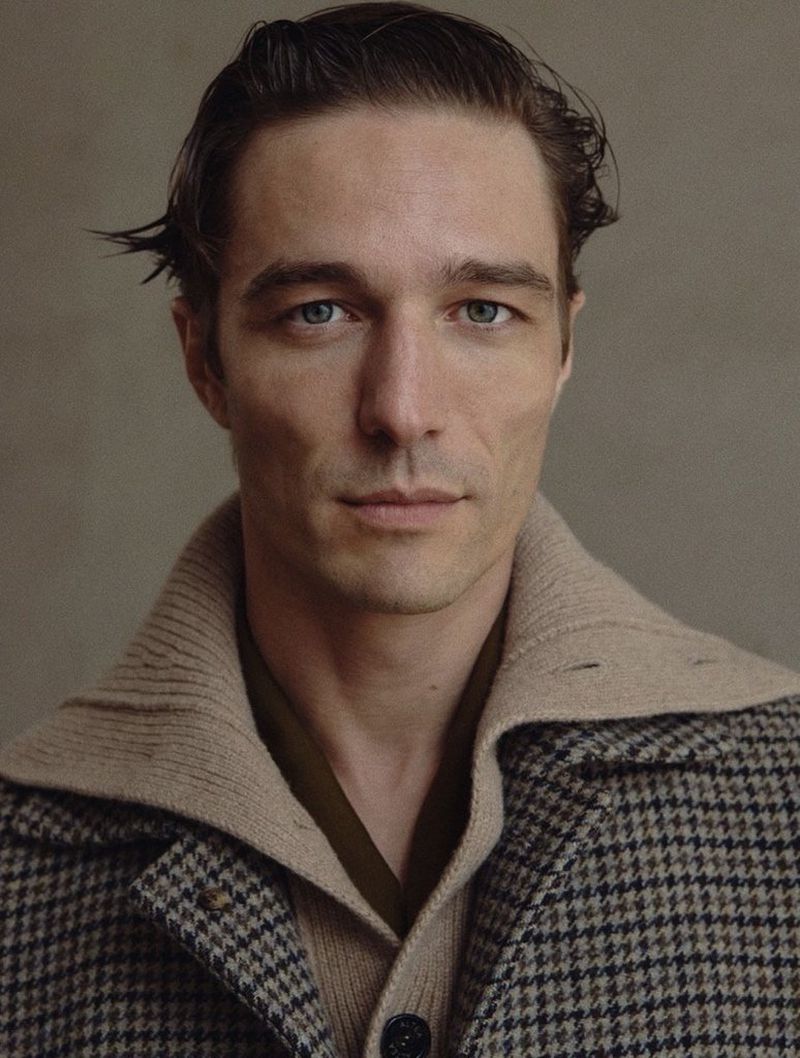 Close-up portrait of a man with blue eyes wearing a patterned coat and tan cardigan, gazing directly at the camera against a neutral background.