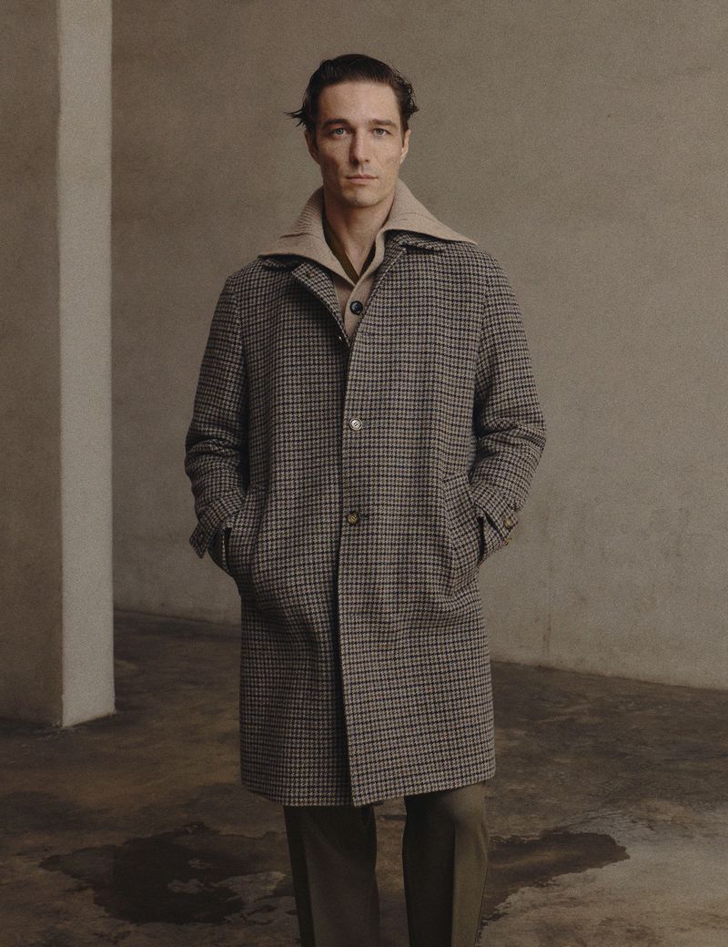 A stylish adult male stands indoors wearing a patterned overcoat, beige collared shirt, and olive dress pants, set against a neutral backdrop.