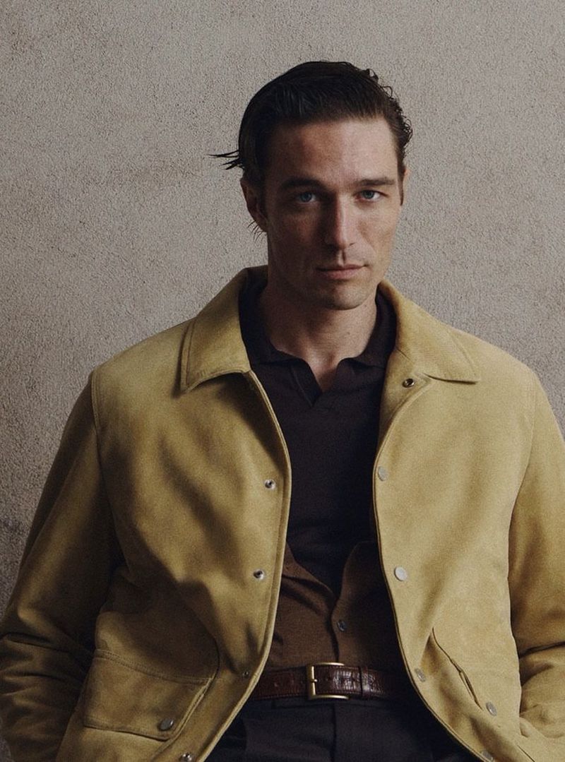 Portrait of a stylish adult male wearing a tan suede jacket and brown polo shirt, set against a neutral grey studio backdrop.
