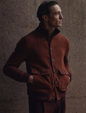 Portrait of a man wearing a suede jacket in front of a textured wall, posing in a contemplative profile view in soft studio lighting.
