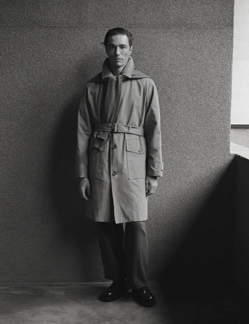 Monochrome portrait of a male model wearing a belted trench coat and sweater over his shoulders against a textured wall.