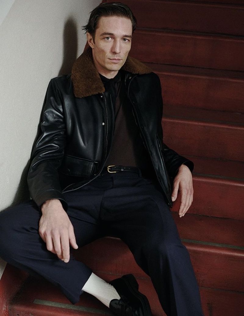 A stylish man in a black leather jacket with a fur collar poses while sitting on red stairs, showcasing an editorial fashion shot.