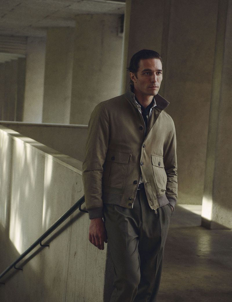 A fashionable adult male poses in a neutral jacket and trousers, set against a concrete interior backdrop with natural lighting.