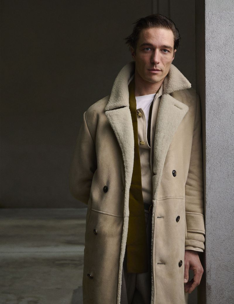 Portrait of a male model in a tan coat standing against a wall with his hands behind his back, looking at the camera with a neutral expression.