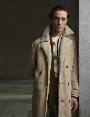 Portrait of a male model in a tan coat standing against a wall with his hands behind his back, looking at the camera with a neutral expression.