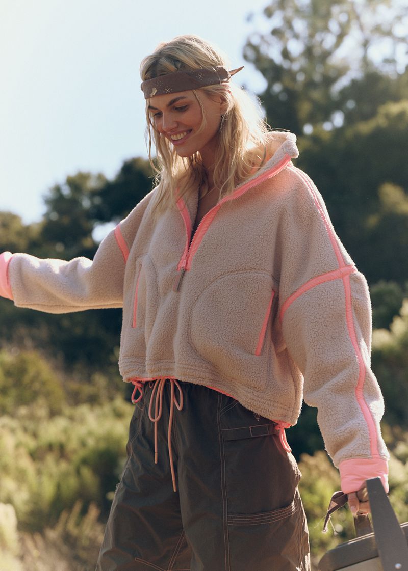 Smiling woman in a sherpa-lined hooded jacket with pink trim poses outdoors in a natural setting with olive-green pants.