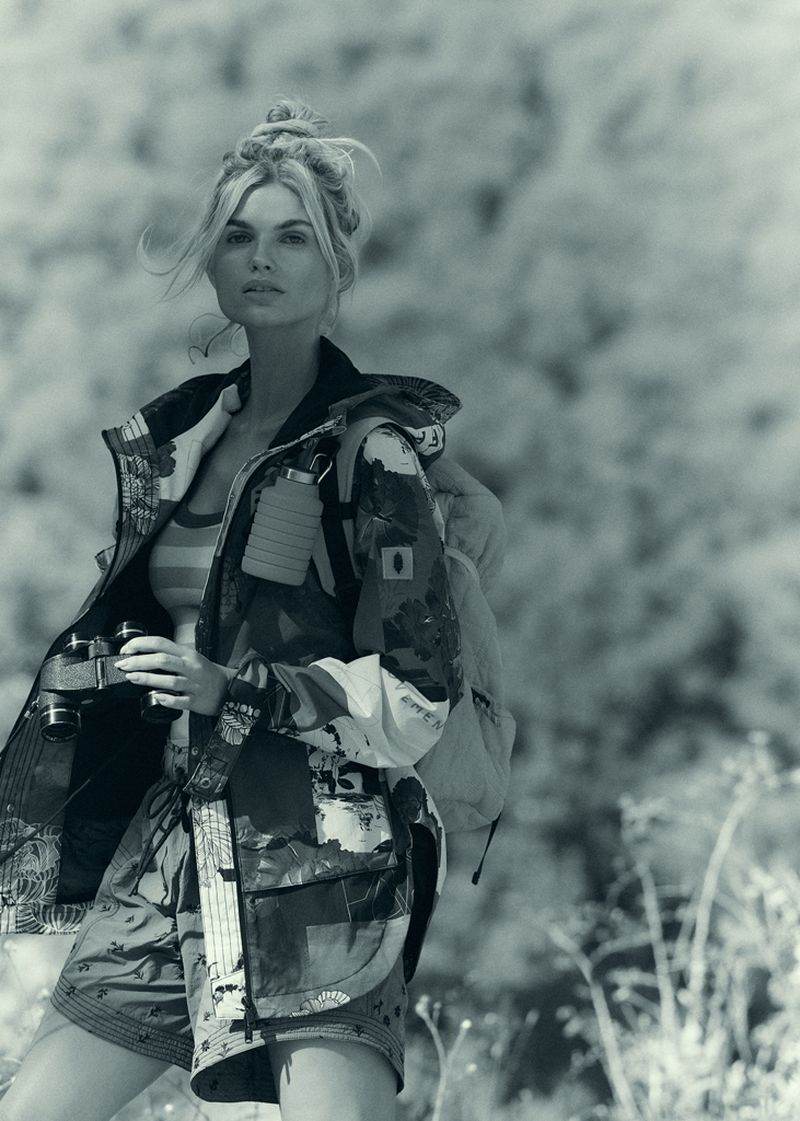 Adult female with blond hair in a messy bun holds binoculars outdoors, wearing a patterned jacket and shorts with a backpack. 