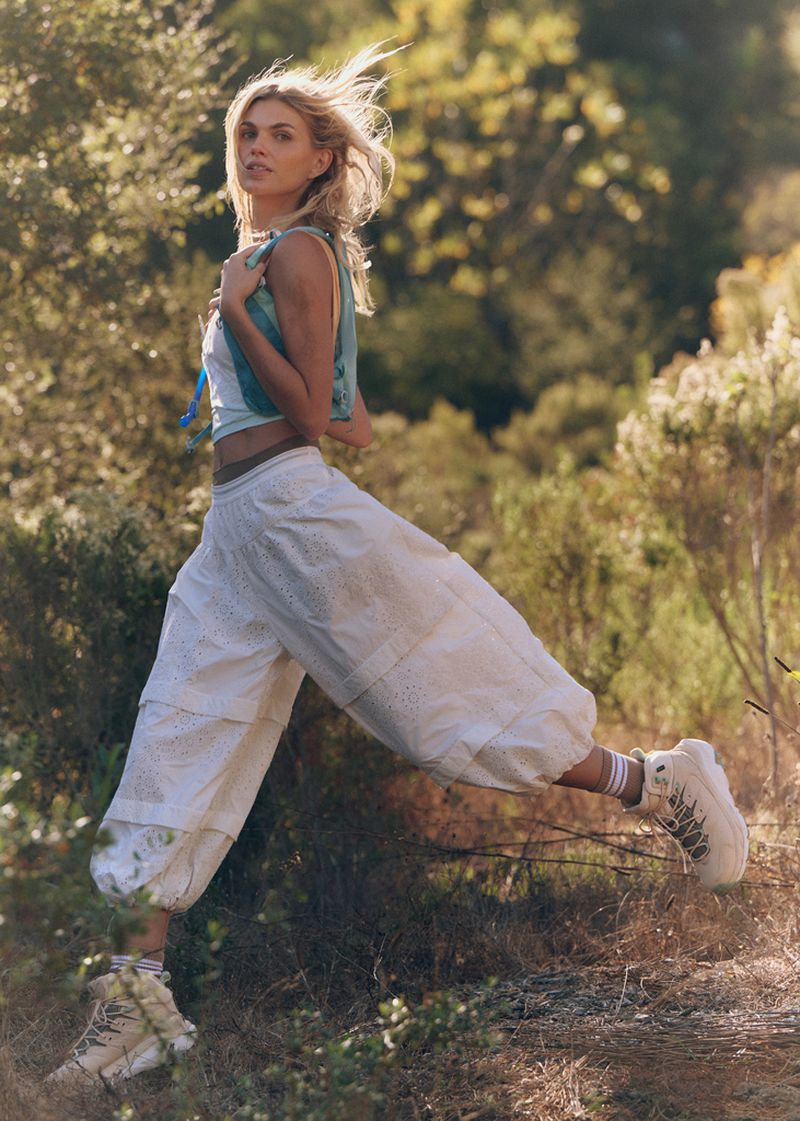 A blonde adult woman in casual summer fashion leaps outdoors in bright sunlight with a forest backdrop. She is wearing a crop top and baggy pants.