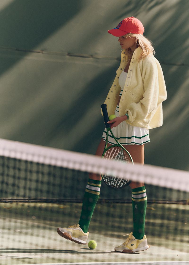 Female tennis player in yellow jacket with tennis racket, ball, pleated mini skirt, tube socks, and sneakers on a tennis court.
