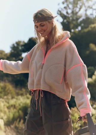 Smiling blonde woman wearing a fleece pullover with pink trim and dark utility pants outdoors in a natural setting.