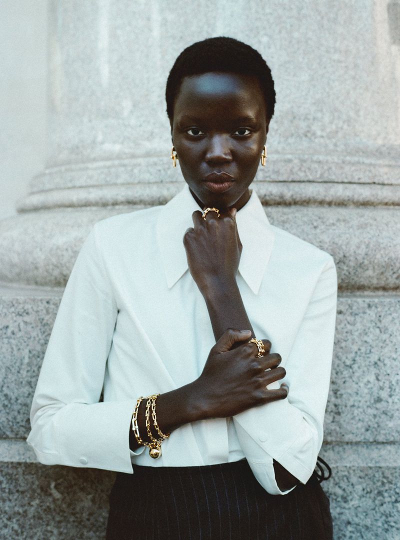 Close-up portrait of a Black woman wearing a white collared shirt and gold jewelry against a stone architectural pillar background