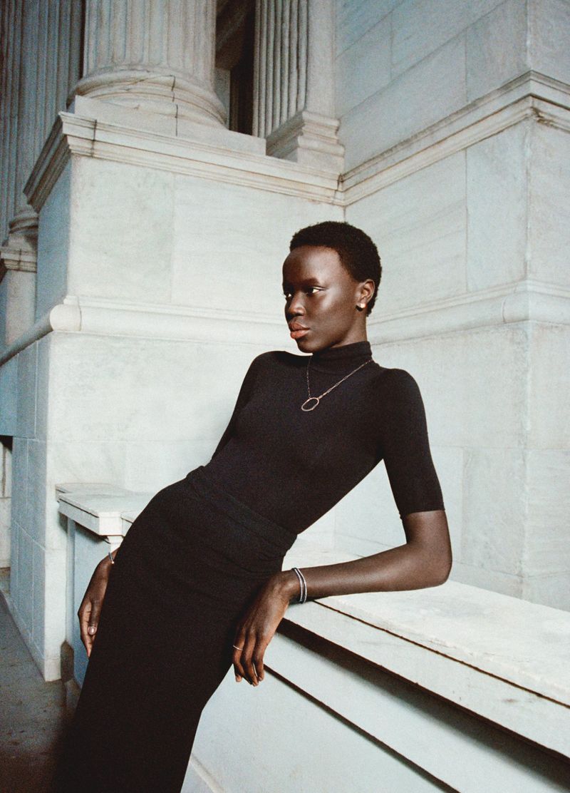 Elegant black woman in black turtleneck dress poses against architectural stone columns, showcasing simple silver pendant necklace and bracelet.