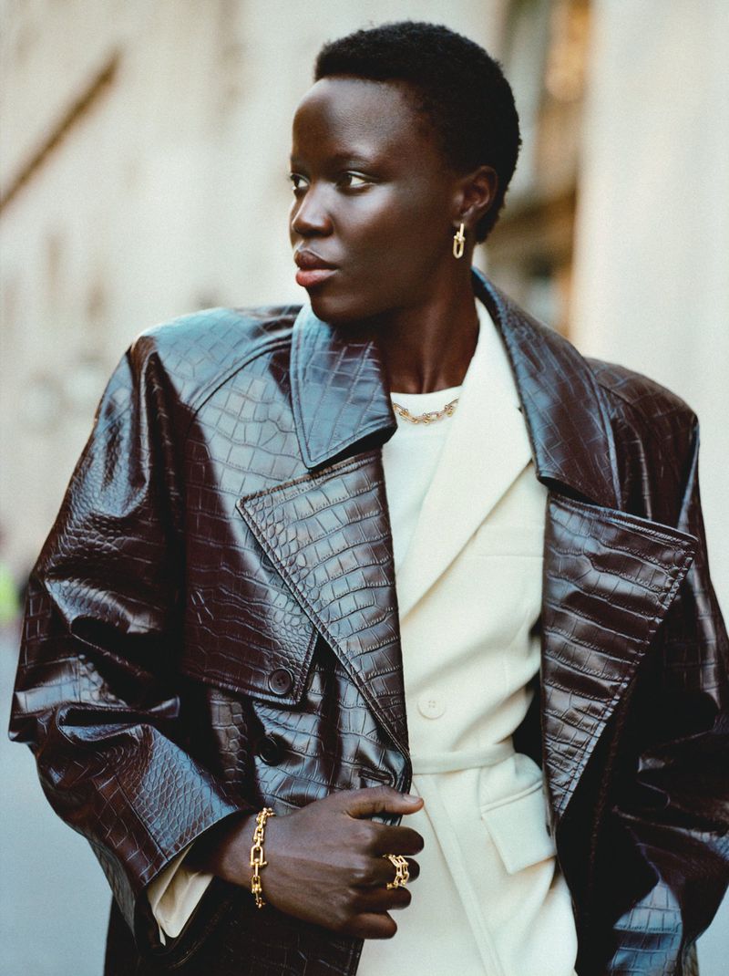 Adult female model poses in a brown, reptile-textured trench coat with jewelry in an outdoor urban setting with neutral tones.